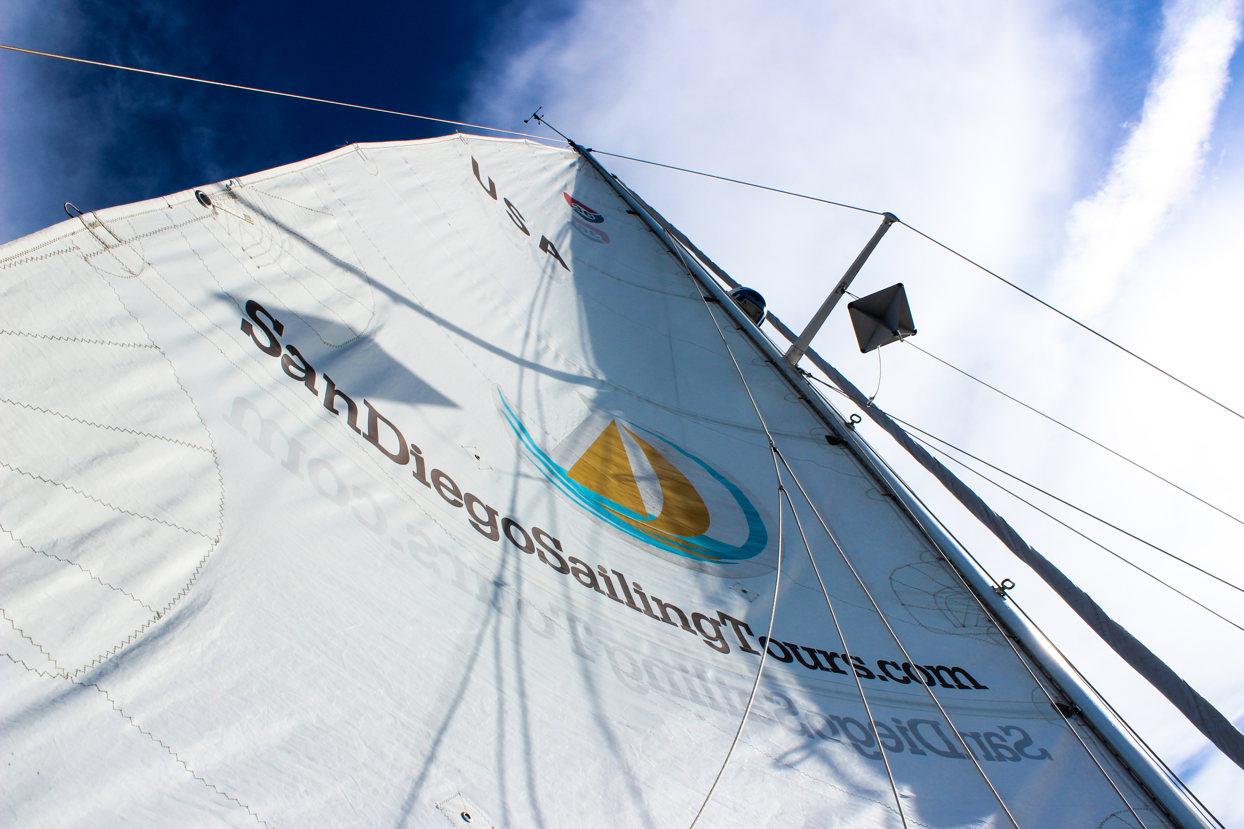 view looking up on a sail with blue skies and clouds above, the sail making a point above viewer. San Diego Sailing Tours logo on sail