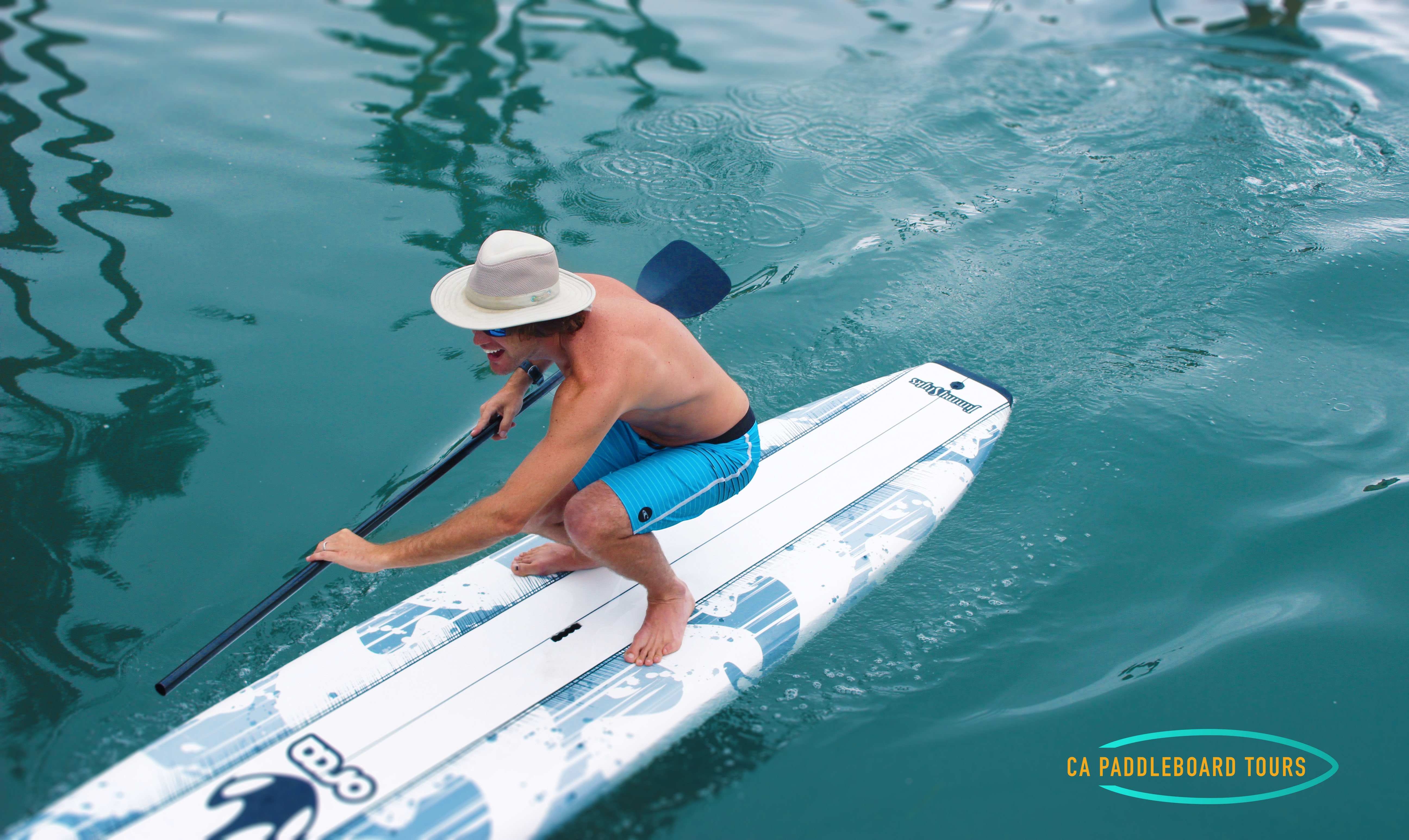 MAN SQUATTING ON PADDLEBOARD AS IT GLIDES IN BLUE WATER.