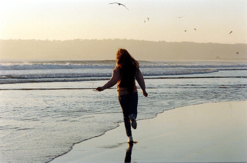 view of girl running, her back turned, along the beach. Point loma in background and the sun is setting with gentle yellow hues.