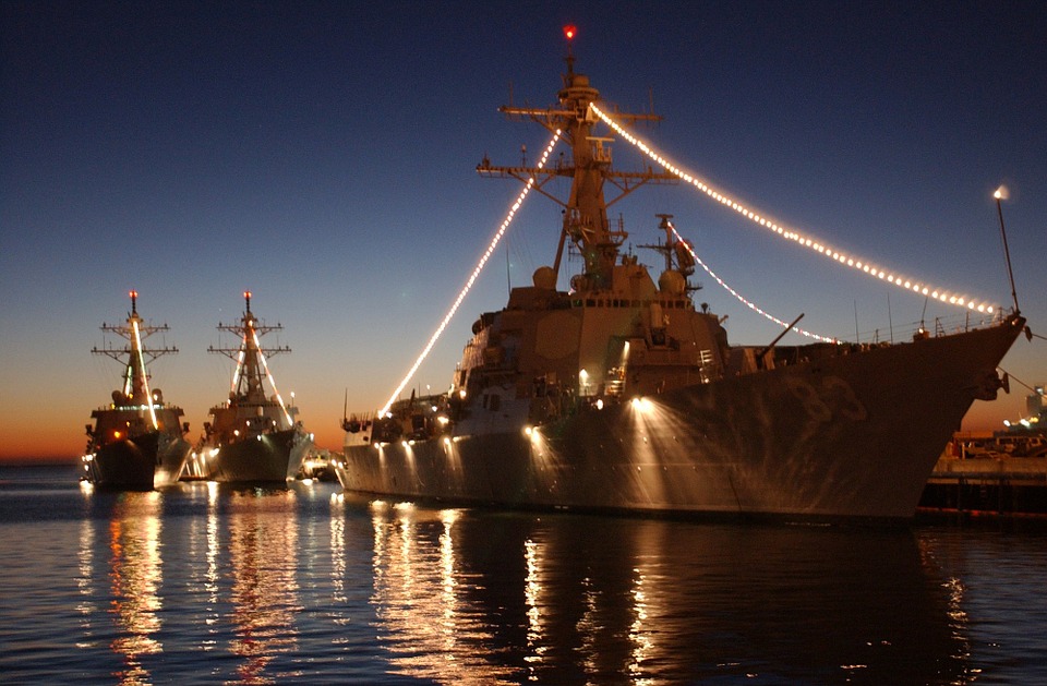 View of 3 large military vessals coming towards viewer from left to right and forward. The sun is almost set, mostly a dark blue, with some red and yellow hues. The ships have lights from bow to mast and along the side, reflecting in water