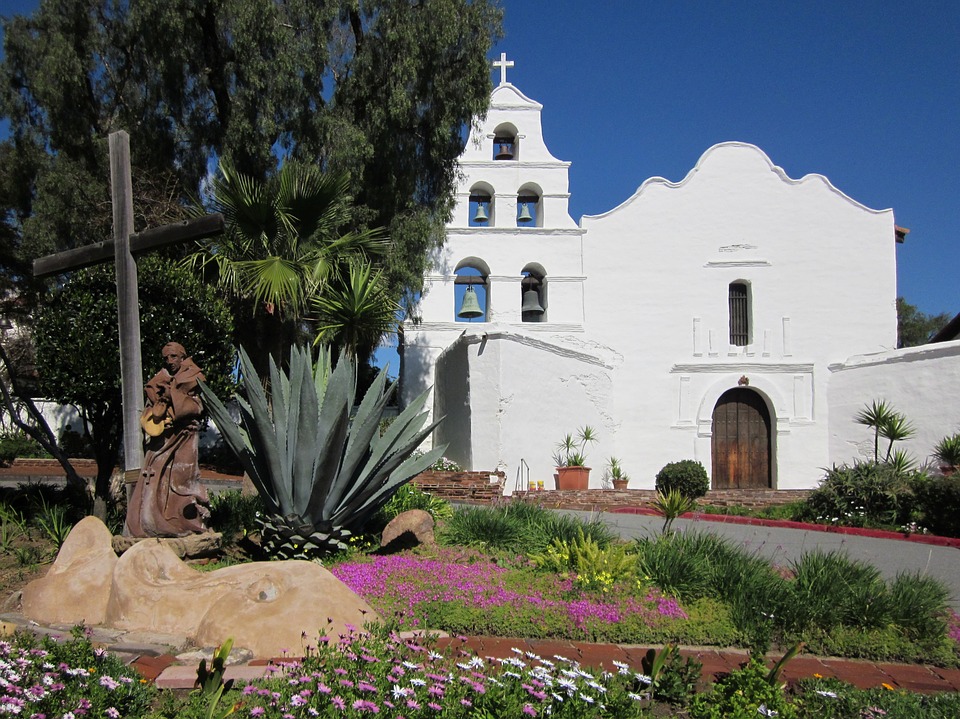 View of San Diego Mission, white adobe with the 5 bells stacked. On the left of the frame is a beautiful native plant garden with blooming flowers, yucca, palm and oak tree, and a wooden cross.